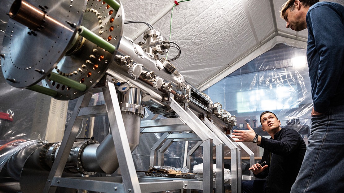 Two engineers working beside a subscale fusion test setup, featuring a long cylindrical chamber with valves, sensors, and piping, installed on a frame inside a controlled lab environment.