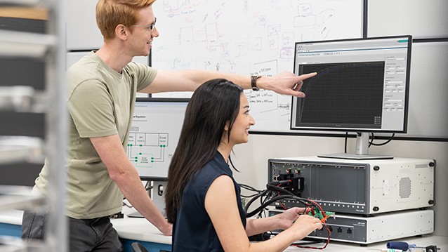 Two people using PXI modular test equipment connected to a computer monitor displaying test software in a lab setting.