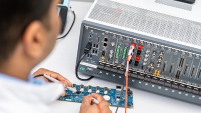 The image shows a person working with electronic equipment, using probes to test or measure a blue circuit board. The board is connected to a PXI system.