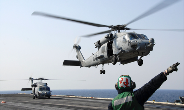 An air traffic controller directs helicopters taking off from an aircraft carrier.