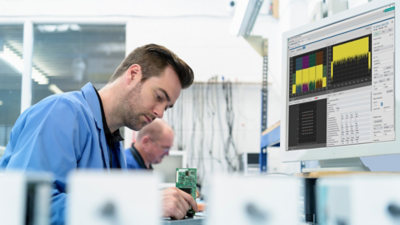 Engineer working at a lab bench, holding a circuit board while monitoring RF measurement data on a computer screen.