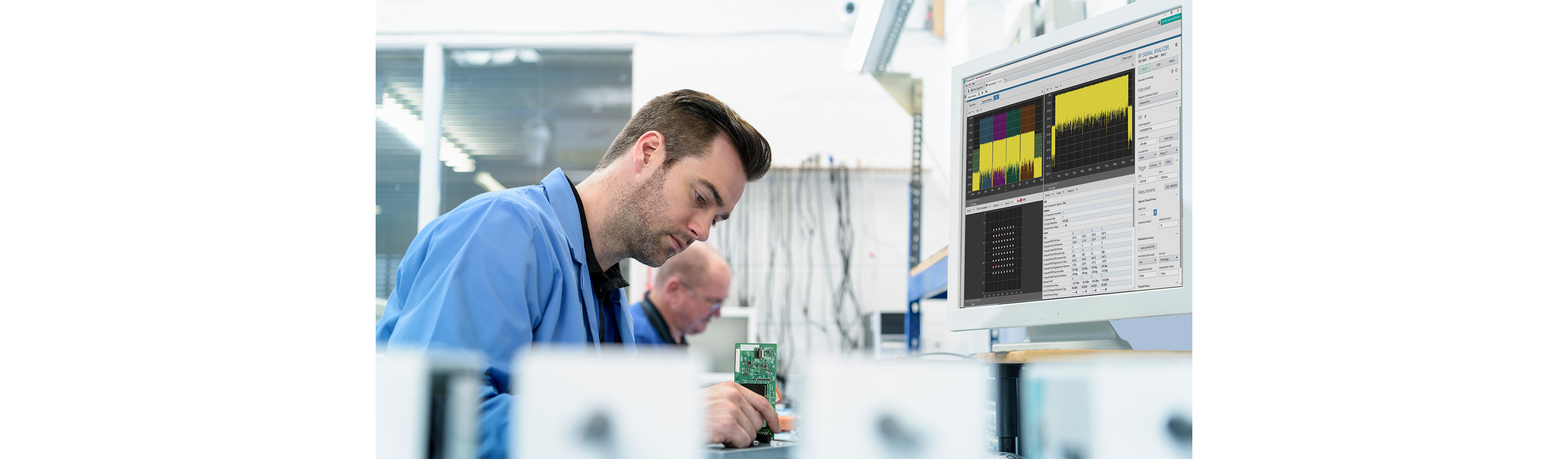 Ingeniero trabajando en un banco de laboratorio, sosteniendo una placa de circuito mientras monitorea los datos de medición de RF en una pantalla de computadora.