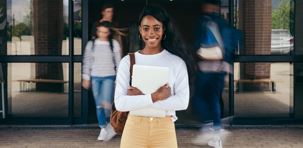 A STEM student stands outside of her school, smiling. Classmates enter and exit the building behind her.