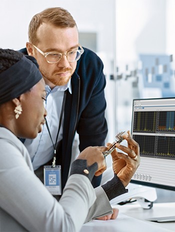 Two engineers in the lab looking over a printed circuit board in front of a computer monitor