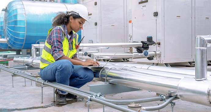 A female engineer in a safety vest working on a laptop and servicing an air conditioner on the rooftop.