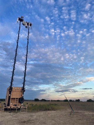 Outdoor communication system featuring two telescopic masts mounted on a trailer, alongside an open control cabinet with circuit boards, wiring, and electronic components for surveillance or signal transmission.