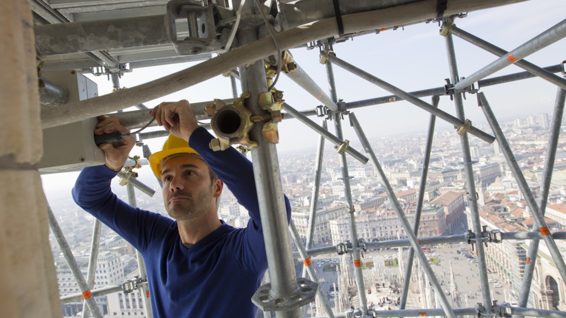 An engineer in a hardhat assembles a strain test system atop a tall building in Milan, Italy.