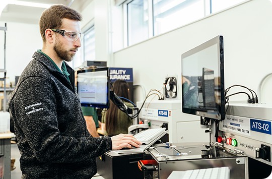 Male engineer in a lab standing in front of a HIL test rack looking at its screen while typing on the keyboard.