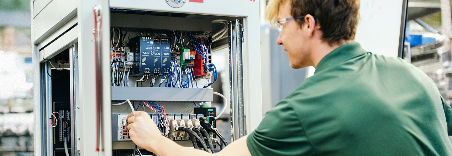 Male engineer in front of an open Hardware-in-the-loop test rack connecting a cable to one of the systems inside the cabinet.    