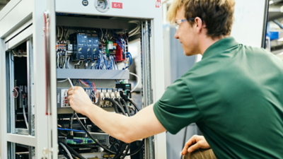 Technician in safety glasses inspecting wiring inside an open electrical control cabinet.
