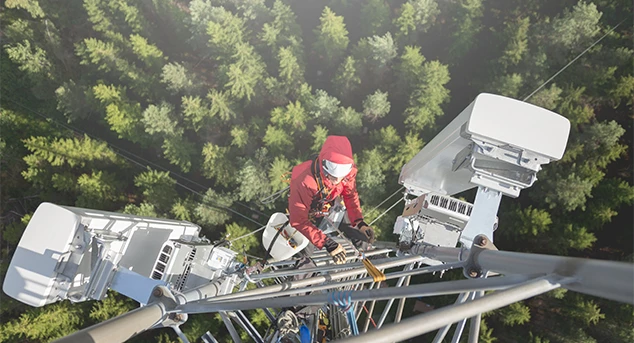 A wireless service technician configures and tests an antenna tower high above a tree-lined road