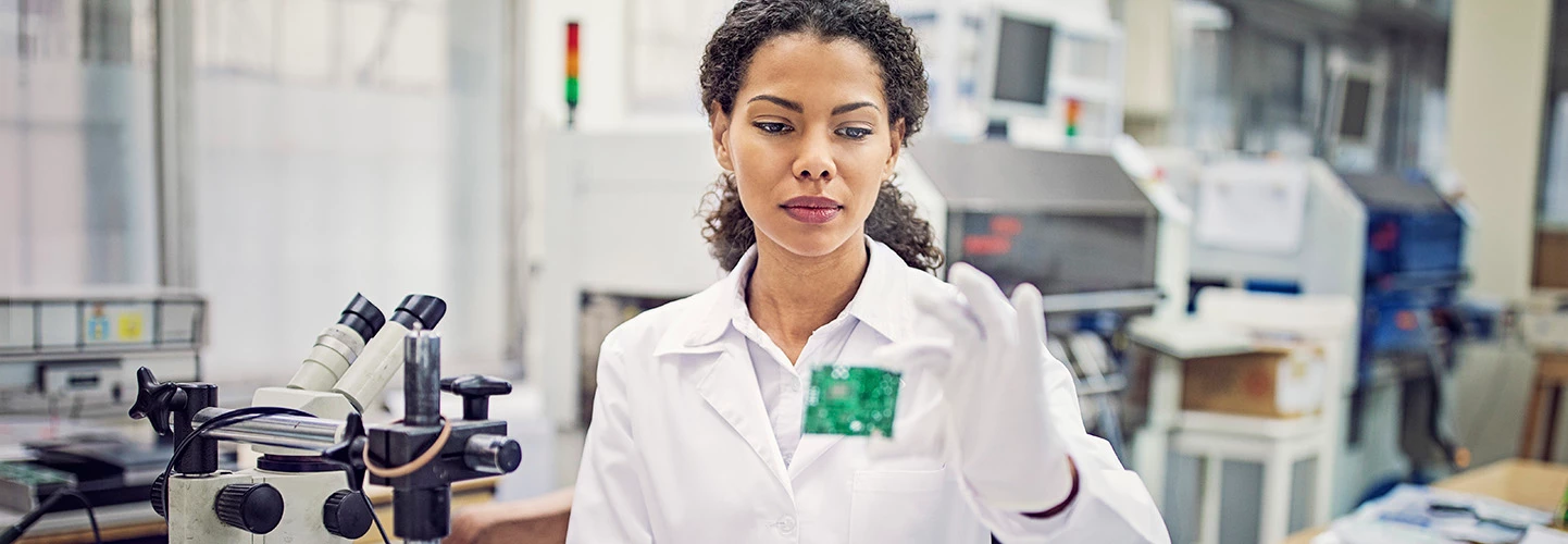 An engineer in a white coat and gloves inspects an integrated circuit board in a modern lab.