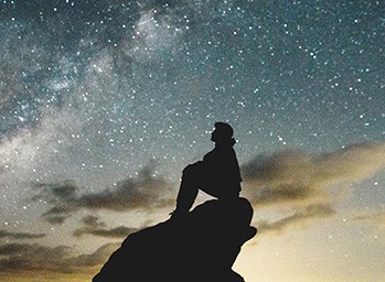 A young person sitting on a hill is in silhouette observing the stars in the night sky