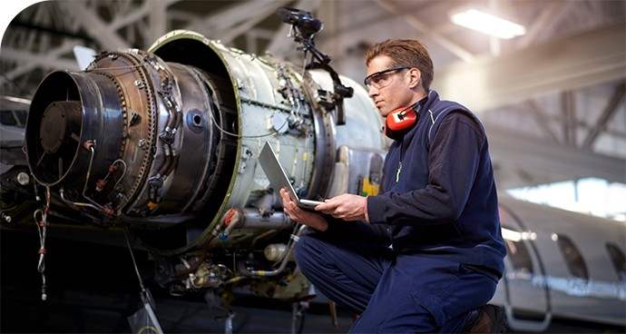 A male engineer kneeling and holding a computer in front of an airplane jet engine getting serviced.