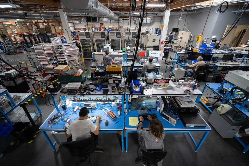 Overhead view of an industrial workshop with technicians assembling and testing electronic components at workstations.