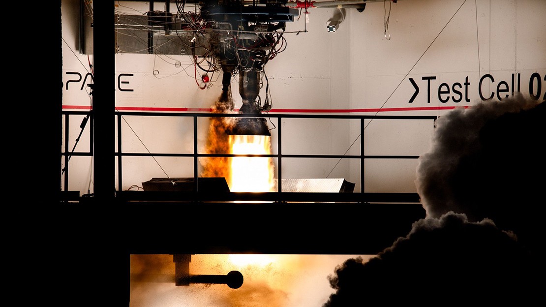 Rocket engine firing during a ground test inside an indoor test facility, with bright flames and exhaust visible beneath the engine, surrounded by metal structures, instrumentation, and partial wall text reading “Test Cell.”