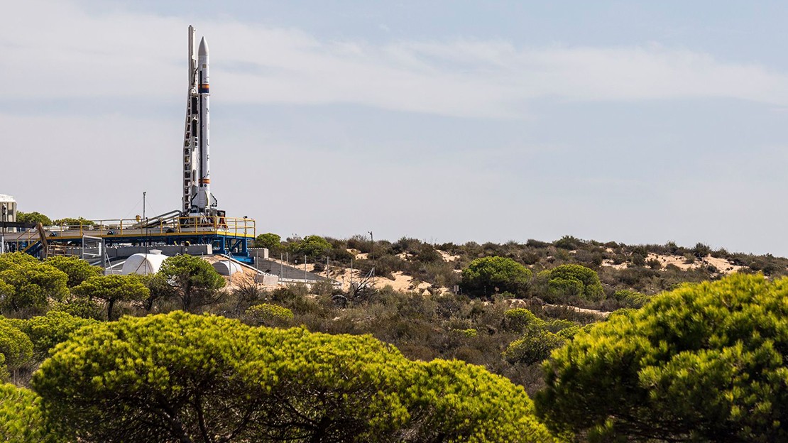 Small rocket standing vertically on a launch pad in a dry, shrub-covered landscape, with metal support structures around the base and a clear sky in the background.