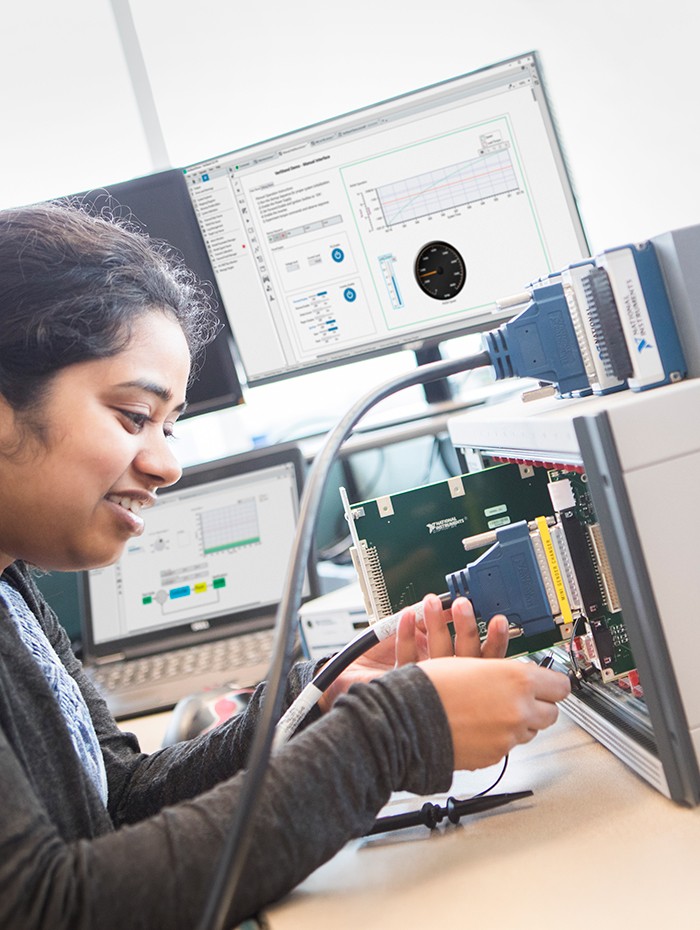 A female engineer connects a cable between a SLSC module and an NI CompactRIO system building up a hardware-in-the-loop (HIL) test system. In the background, NI VeriStand software is monitoring another test on nearby computer screens.​ 