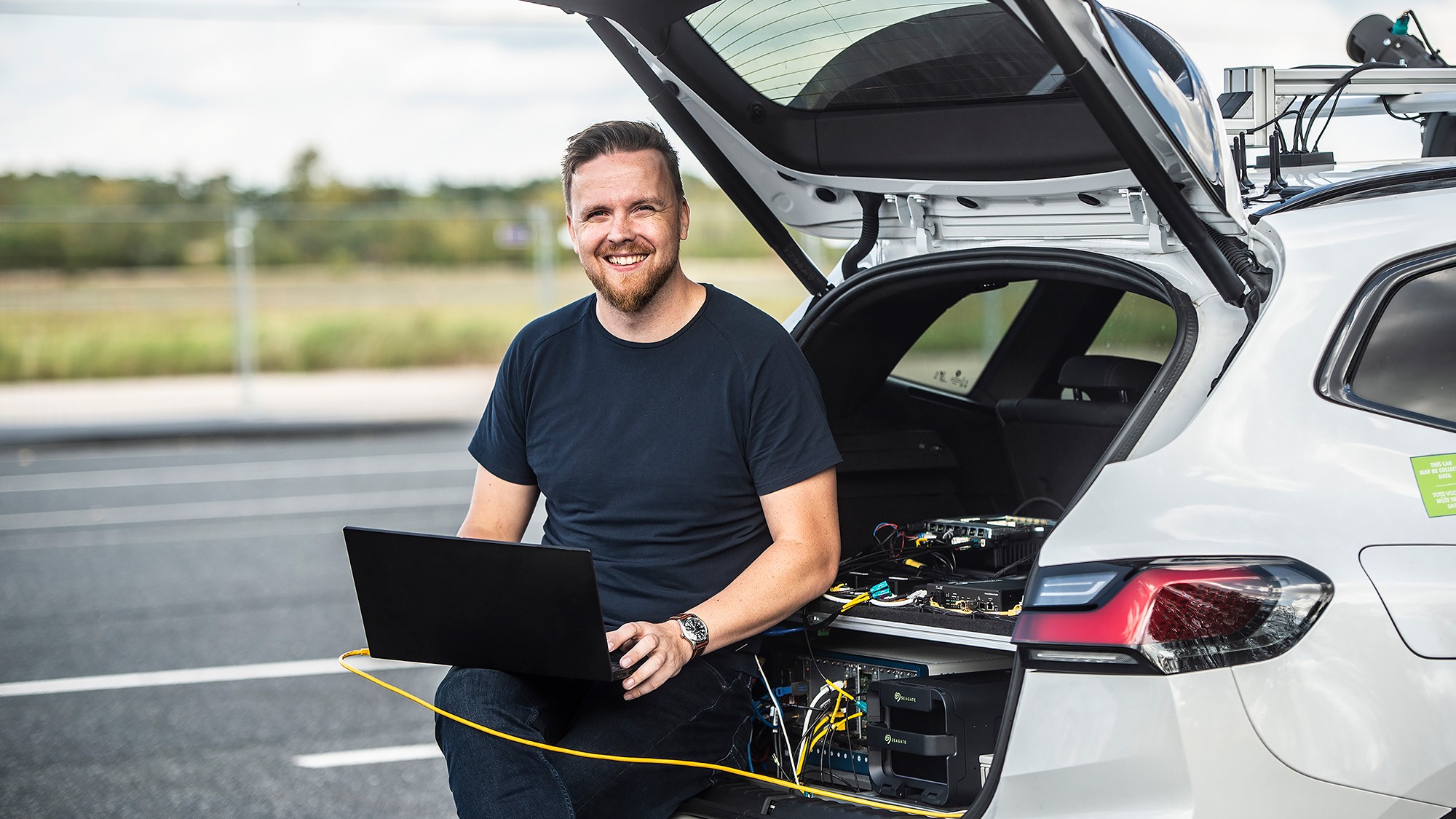 Engineer stands beside a vehicle with the rear hatch open, using a laptop connected to ADAS validation and data acquisition equipment.
