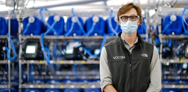 Sam Burner, manufacturing engineer at Ventec Life Systems, stands in front of shelves full of ventilators. He is wearing a face mask and eye protection.