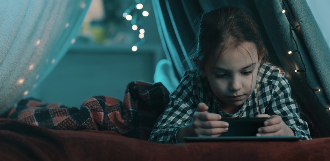 A child watches videos on her phone in a blanket fort decorated with LED string lights.