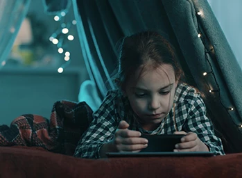 A child watches videos on her phone in a blanket fort decorated with LED string lights.