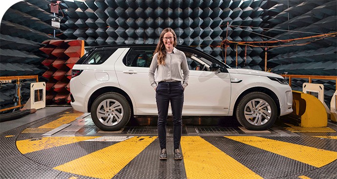 A female engineer standing in front of a white car on a dynamometer within an anechoic chamber.